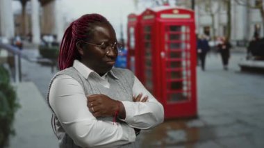 Şehrin arka planında kollarını kavuşturmuş kırmızı telefon kulübesi olan bir kadın açık şehir ortamını öneriyor..
