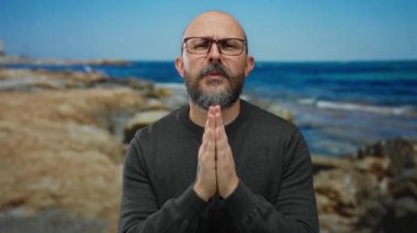 Deniz kenarında gözlüklü bir adam, güneşli bir günde elleri ile okyanusa doğru hareket eder, sakin bir sahil ortamında duygularını ve düşüncelerini aktarır..