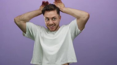 Sakallı genç bir adam, mor bir arka planda, elleri başının üstünde, eğlenceli, kaprisli bir ifade ve neşe sahnesi yaratıyor.