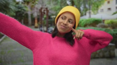 Pembe kazaklı ve sarı bereli İspanyol kadın canlı yeşil bir parkta güven içinde poz veriyor, neşeli bir açık hava havası sergiliyor..