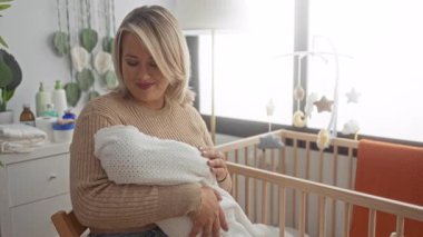 Stüdyoda ahşap beşiğin yanında beyaz battaniyeye sarılı uyuyan bebeği nazikçe kucaklar; anne sevgisi dinginliği.