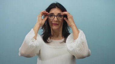 Kadın, mavi stüdyo ortamında iki eliyle gözlüğü çıkarırken doğrudan ileriye bakıyor; kendine güven netliği odaklama görüşü.