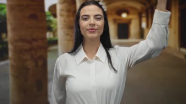 Beyaz tişörtlü genç İspanyol kadın eski bir üniversite avlusunda gülümserken eliyle gözlerini kapatıyor..