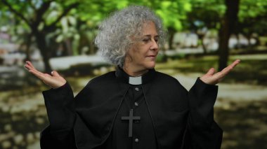 Gri saçlı, papaz kıyafeti giymiş yaşlı bir kadın sakin bir açık hava parkı ortamında duruyor. Dalgınlığı ve maneviyatı yemyeşil ağaçların altında somutlaştırıyor..