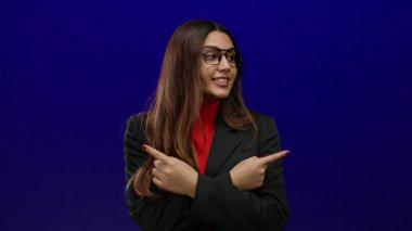 Gözlüklü ve siyah takım elbiseli İspanyol kadın canlı mavi arka plana karşı güvenle yanlamasına bakıyor, yönünü ve kararını odaklanmış bir ifadeyle gösteriyor..