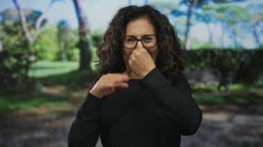 Gözlüklü İspanyol orta yaşlı kadın yeşil park ortamında hoşnutsuzluğa tepki veriyor. Gündüz vakti çeşitli duygular ve vücut dilleri sergiliyor..