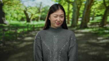 Güneşli bir günde yemyeşil bir parkta neşeyle göz kırpan uzun saçlı ve gülümseyen bir kadın.