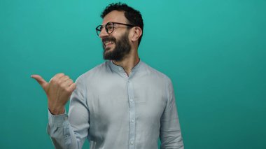 Sakallı İspanyol adam gülümsüyor ve yeşil arka planda parmağıyla işaret ediyor açık gömlek ve gözlük takıyor..