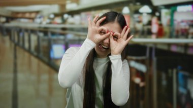 Uzun saçlı, gözlüklü, elleri açık alışveriş merkezinin içinde beyaz gömlekli, hareketli atmosfer ve canlı mağazalarla çevrili bir kadın.