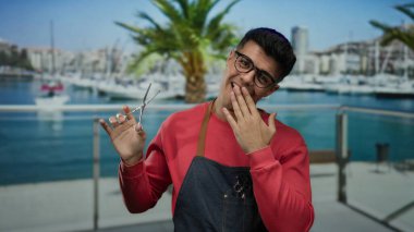 Deniz kıyısında bir limanda elinde makasla dolaşan gözlüklü ve önlüklü genç adam bir tekne ve palmiye ağacına karşı neşeli bir ifade sergiliyor..