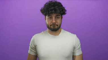 Beyaz tişörtlü, sakallı, genç İspanyol adam canlı mor arka plana karşı duruyor. Düşünceli bir yüz ifadesi var. Modern gündelik stil ve izolasyon kavramı sergiliyor.