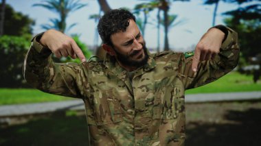 Sakallı, askeri üniformalı İspanyol bir adam arka planda ağaçlarla güneşli yeşil bir parkın aşağısına işaret ediyor..