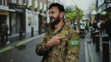 Sakallı, kamuflaj üniformalı bir asker Brezilya bayrak kuşağını işaret ediyor açık bir şehir caddesinde hareketli duruyor..