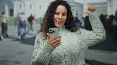 Genç İspanyol bir kadın, turistlerle dolu kalabalık bir caddenin ikonik kulesinin yanında, açık havada akıllı bir telefonla selfie çekiyor..