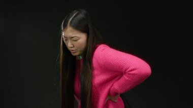 Pembe kazaklı kadın, izole edilmiş siyah arkaplan karşısında acı çekiyor. Rahatsızlık ve duruş sorunu olduğunu gösteriyor..