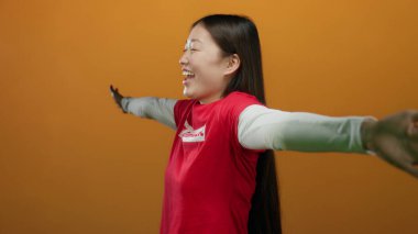 Turuncu bir duvarın önünde kollarını açmış gülümseyen bir kadın, mutluluk ve özgürlük yayıyor, modern ve izole bir ortamda mutluluk, pozitiflik ve canlılığı somutlaştırıyor..