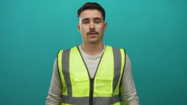 Yansıtıcı yelek giyen genç İspanyol adam, izole edilmiş yeşil duvar arka planına karşı duruyor. Düşünceli ve sakin görünüyor..