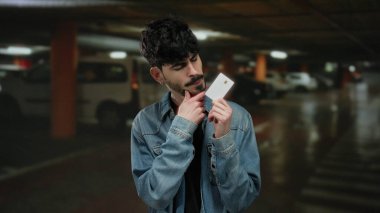 Kot ceketli genç adam loş ışıklı bir park alanında bir kart tutuyor, dikkatlice bakıyor, tefekkür etmeyi ya da karar vermeyi öneriyor..