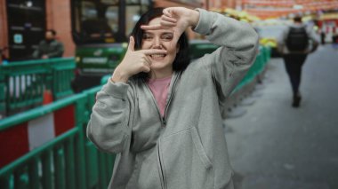 Caddede elleri gri kapüşonlu bir kadın jesti yapıyor. Şehir ortamında neşeli bir ifade sergiliyor. Yayalar ve renkli barikatlar kuruyor..