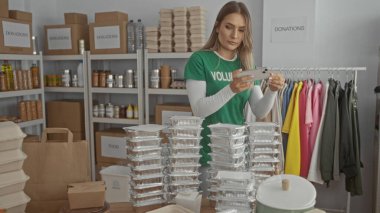 Bağış kutuları, yiyecek konteynırları ve giysi rafları ile çevrili hayır kurumunda akıllı telefon kullanan gönüllü kadın, iç mekan ve toplum yardımını vurguladı.