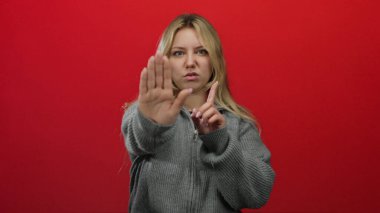 Kadın jestleri canlı kırmızı bir arkaplanda duruyor, ciddi bir ifade sergiliyor ve gri bir kazak giyiyor, hoşnutsuzluk ve kendine güvensizliği vurguluyor..