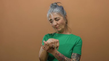 Yeşil tişörtlü, gri saçlı yaşlı kadın ellerini izole edilmiş kahverengi arka plan duvarına doğru inceliyor..
