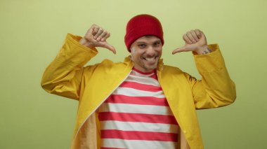 Sarı yağmurluklu ve kırmızı bereli İspanyol genç adam sarı arka planda kendini işaret ediyor. Kendine güveni ve kendine güveni var ve gülümsüyor..