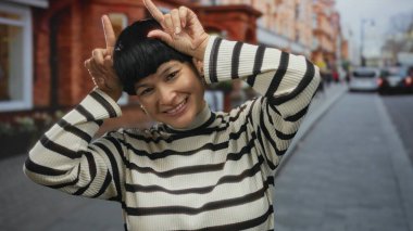 Genç Çinli bir kadın, şehir ortamında çizgili bir süveter giyerek, kalabalık bir caddede gülümseyerek elleri başının üstünde şakacı boynuzlar sallıyor..