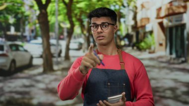Kırmızı kazak ve kot önlük giyen İspanyol genç adam ağaçlarla ve park halindeki arabalarla dolu bir sokakta kalemle notlar alıyor.
