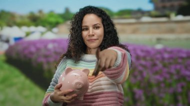 Parkta elinde domuz kumbarası olan genç İspanyol kadın kameraya gülümsüyor ve arka planda canlı çiçekler ve yeşillikle işaret ediyor..