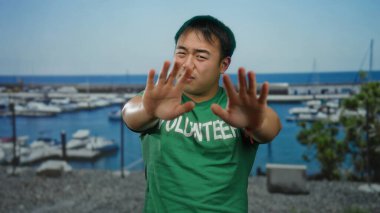 Üzerinde gönüllü yazan yeşil gömlekli genç adam açık mavi gökyüzünün altında bulanık teknelerle sahil limanında yüzünü kapatıyor.