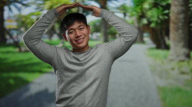 Parktaki genç adam canlı yeşil ağaçlar ve açık mavi gökyüzü arkaplanı arasında neşeli jest yapıyor. Rahatlamış açık hava atmosferi ve neşeli bir ifade sergiliyor.