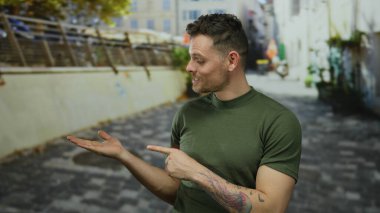 Yeşil gömlekli genç adam şehir caddesinde duruyor. Dövmesi görünür, canlı şehir zeminiyle çevrili, odaklanmış bir ifadeyle modern şehir yaşamını gözler önüne seriyor..