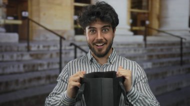 Dışarıda elinde hediye çantasıyla gezen genç adam sürpriz ya da kutlama beklentisiyle gülümsüyor..