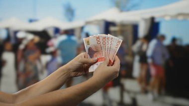 Norveç kronu taşıyan bir adam açık pazar caddesinde, çeşitli tezgah ve dükkanların yakınında alışveriş yapan insanlarla çevrili..