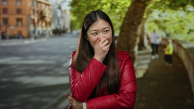 Kırmızı ceketli kadın, arka planda ağaçlar ve binalarla şehir caddesinde gülümsüyor.