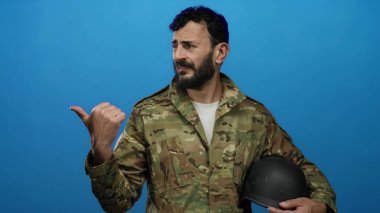 Mavi arka planda miğfer tutan askeri üniformalı İspanyol sakallı adam düşünceli bir yüz ifadesi sergiliyor ve başparmağıyla işaret ediyor..