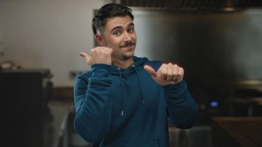 Mavi kapüşonlu genç adam gülümsüyor ve parmaklarını mutfakta yana doğru doğrultuyor. İçeriden canlı bir atmosfer ve dostça bir jest sunuyor..