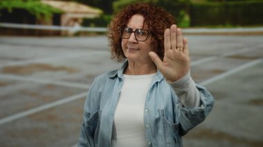 Açık tenis kortunda el kol hareketi yapan, gözlüklü ve mavi gömlekli bulanık yeşilliklerle çevrili bir kadın..