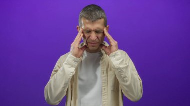 Kısa saçlı, bej gömlekli bir adam, mor bir arka plan duvarına karşı düşünceleriyle şakaklarına dokunur, stres ve düşünce duyguları taşır..