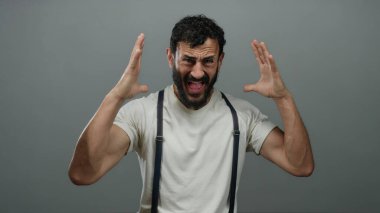 Sakallı İspanyol bir adam beyaz gömlekli, pantolon askısı takmış gri bir arka planda hayal kırıklığı yaşıyor..