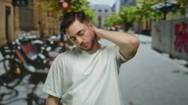Sakallı genç adam canlı bir şehir caddesinde duruyor, canlı yeşillik ve şehir manzarası ile çevrili, modern açık hava yaşamını yansıtıyor..