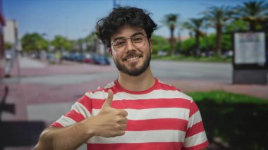 Sakallı, dışarıda gülümseyen ve arka planda palmiye ağaçları ve arabaları olan bir sokakta kendini işaret eden Latin asıllı genç adam..