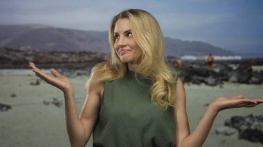 Kadın kollarını arka planda dağlar olan manzaralı bir plajda açıyor, sakin bir açık hava manzarası sergiliyor, gülümsemesi ve rahat bir duruşu var..