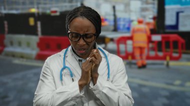 İnşaat alanındaki doktor laboratuvar önlüğü ve steteskop giyip dışarıda el sıkışık bir şekilde dikiliyor..