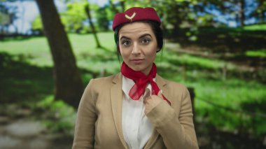 Hostes üniformalı bir kadın, güneşli bir parkta kırmızı bir eşarp ve şapka takarak jest yapıyor. Yeşilliğin ortasında düşünceli ve anlamlı bir duruş sergiliyor..