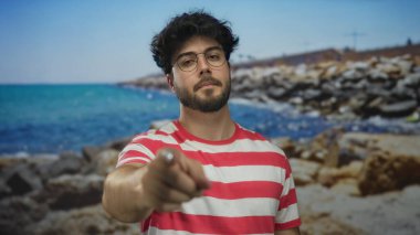 Sakallı, gözlüklü, çizgili tişörtlü genç adam kumlu sahilde, açık gökyüzünü işaret ederek kıyıda duruyor..