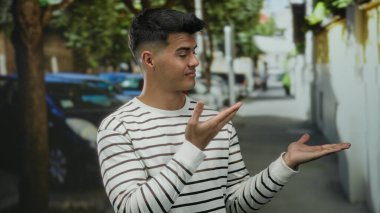 Çizgili gömlekli genç adam, şehir arka planında bir sokak ve park etmiş arabalarla avucuna doğru hareketler yapıyor, açık havada kendine güvenen ve rahat bir poz veriyor..
