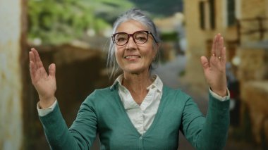 Gri saçlı, gözlüklü yaşlı bir kadın canlı bir sokakta jestler yapıyor. Arka planda bulanık binalar ve yemyeşil alanlar var. Açık hava kentsel ortamı öneriyor..