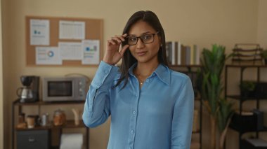 Gözlüklü genç Latin kadın, kitaplar ve bitkilerle çevrili modern ofis ortamında güvenle duruyor, profesyonelliği ve odaklanmayı aktarıyor..